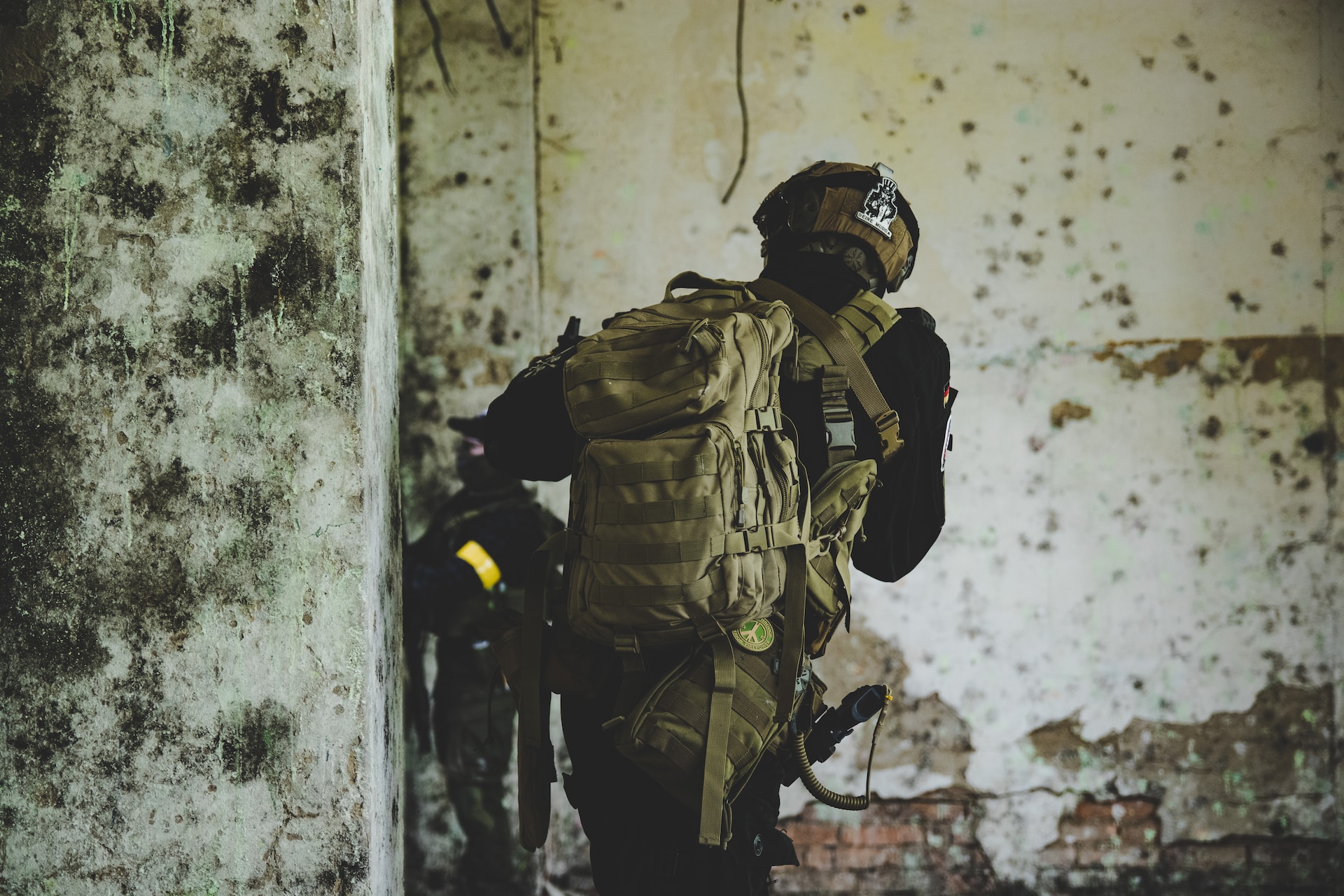 A soldier in tactical gear and a helmet, carrying a large backpack, moves cautiously through a dilapidated, moldy building—an environment ideal for experiential simulations. Another soldier is partially visible in the background.