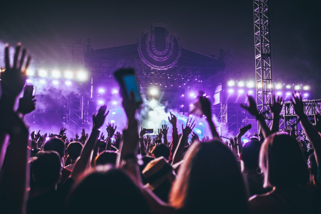 A crowd of people with raised hands enjoying a nighttime music concert, brightly lit by colorful stage lights, with a large stage and electronic display in the background.