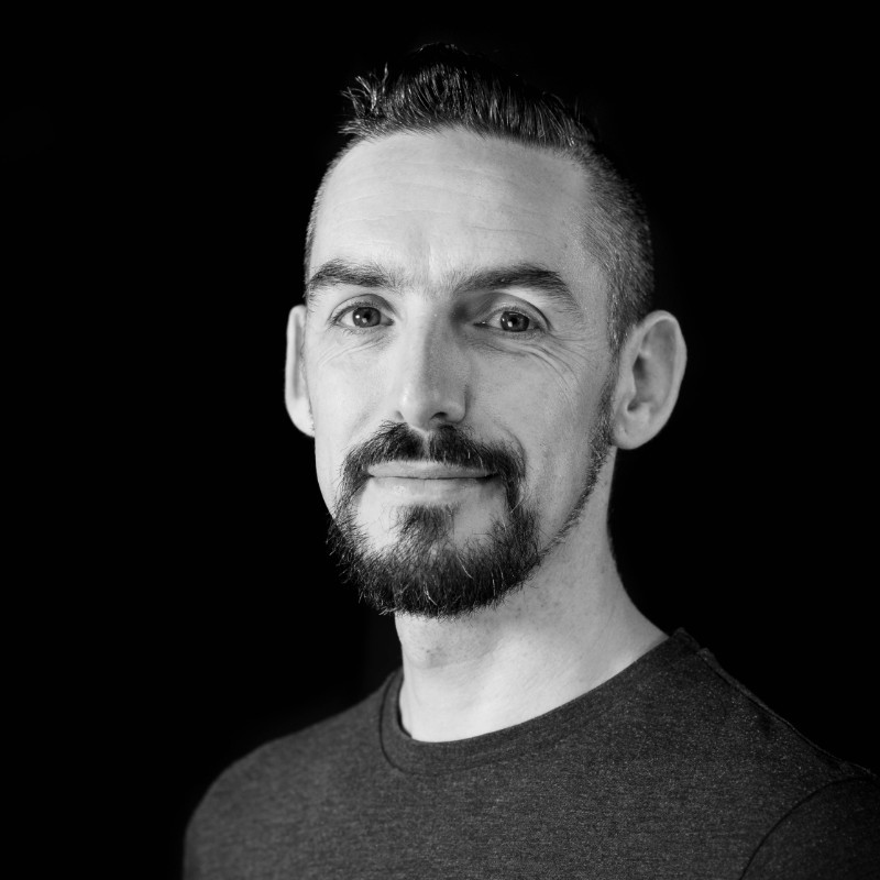 Black and white portrait of a man with short dark hair and a trimmed beard, wearing a plain dark t-shirt, smiling slightly—perfect for sharing marketing insights—set against a black background.