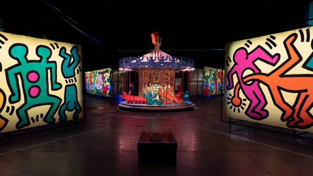 A brightly lit carousel is surrounded by large panels featuring colorful, bold, outlined figures in the style of Keith Haring, evoking the creative energy often found in brand homes, displayed in a dark indoor exhibition space.