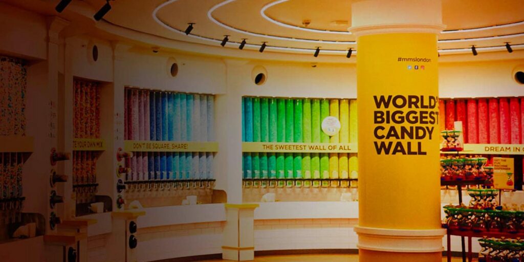 A brightly lit store features a large yellow pillar that reads WORLD BIGGEST CANDY WALL. Behind it, rows of colorful candy dispensers are arranged by color along the curved wall.