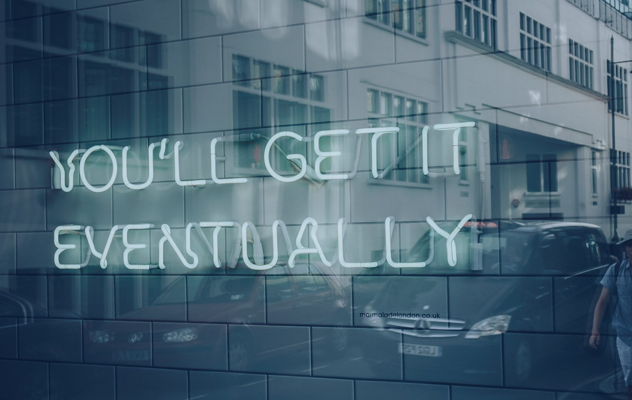 Neon sign on a tiled wall reads YOULL GET IT EVENTUALLY; building windows and parked cars are reflected in the glass, creating an experiential marketing vibe that hints at building loyalty over time.