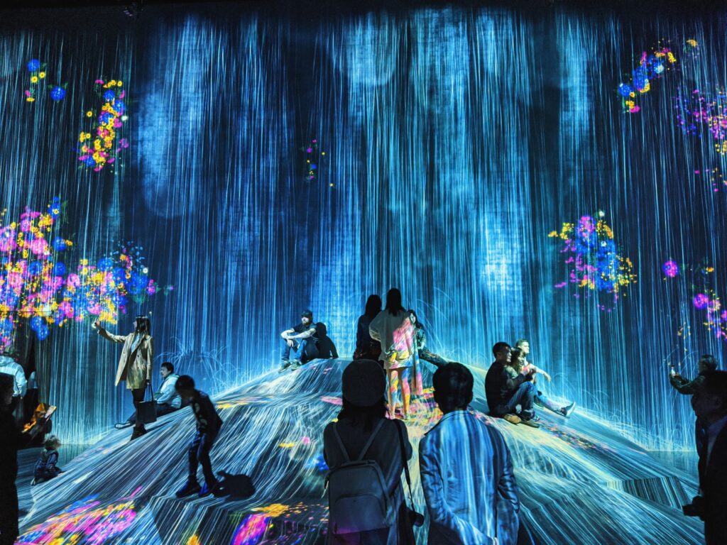 People interact with a digital art installation featuring cascading blue lights and colorful projected flowers, creating an immersive, futuristic environment. Some individuals stand, sit, or pose on a central mound under the lights.