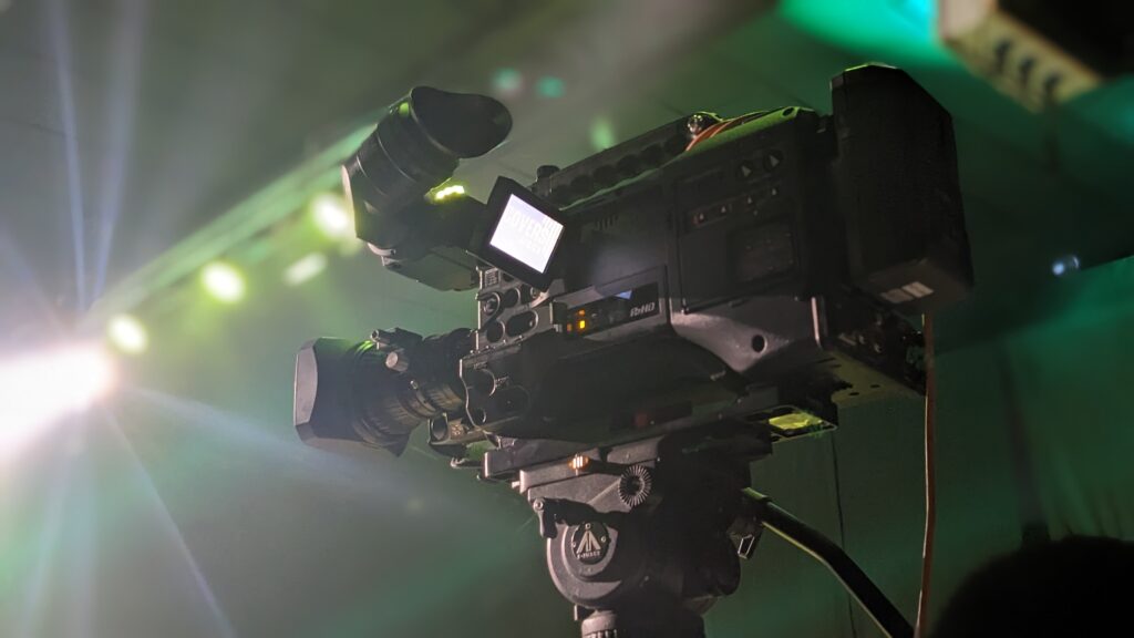 A professional video camera on a tripod is set up in a dimly lit environment with green lighting and lens flare in the background.