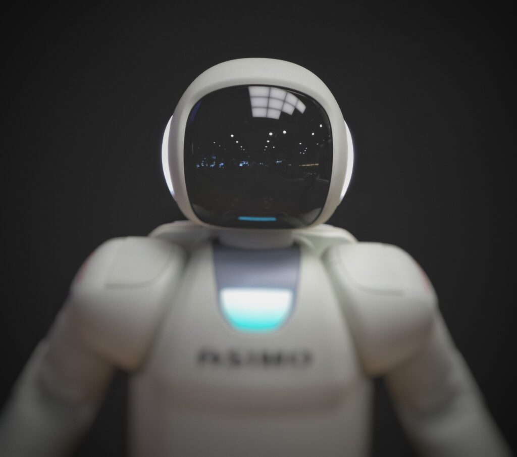 A white humanoid robot with a shiny black faceplate and glowing blue chest light stands against a dark background. Reflections of lights are visible on its face.