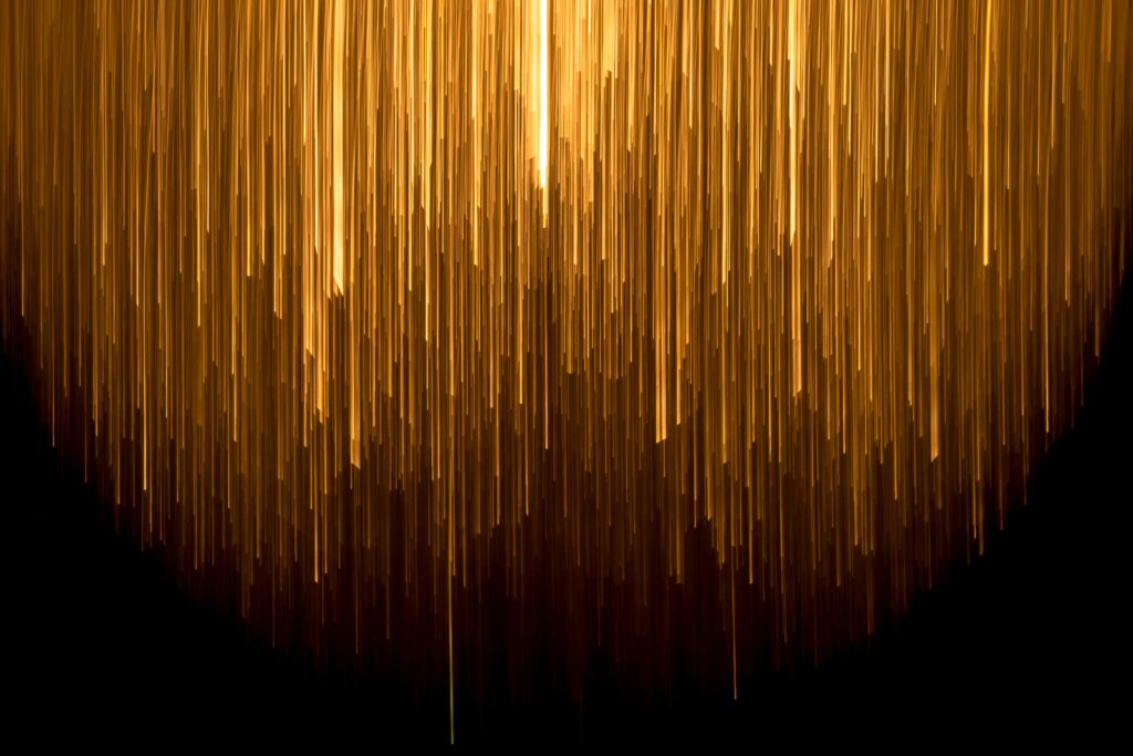 Vertical streaks of golden light cascade downward against a black background, creating an abstract, glowing, rainfall-like effect.