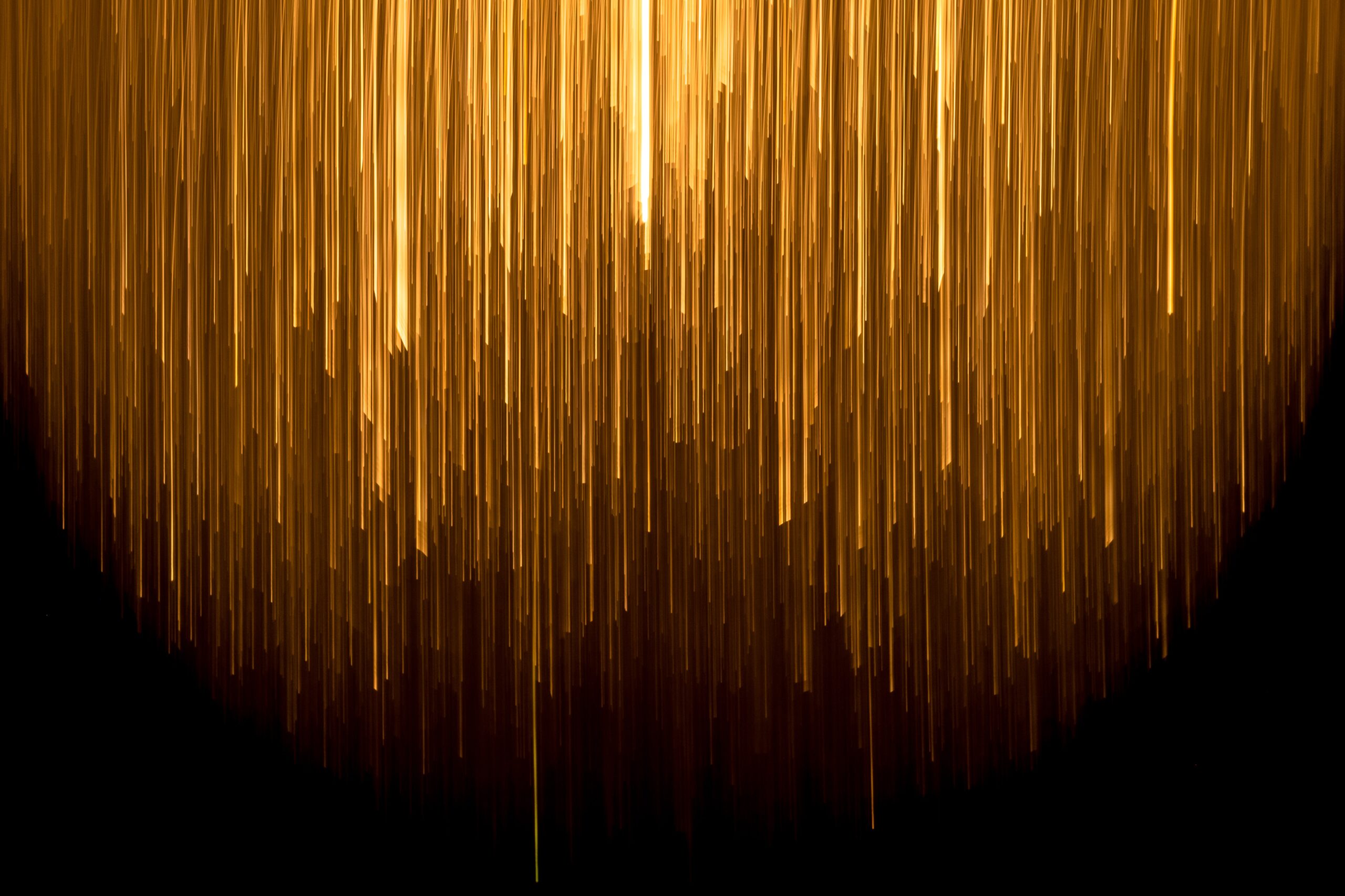 Vertical streaks of golden light cascade downward against a black background, creating an abstract, glowing, rainfall-like effect.
