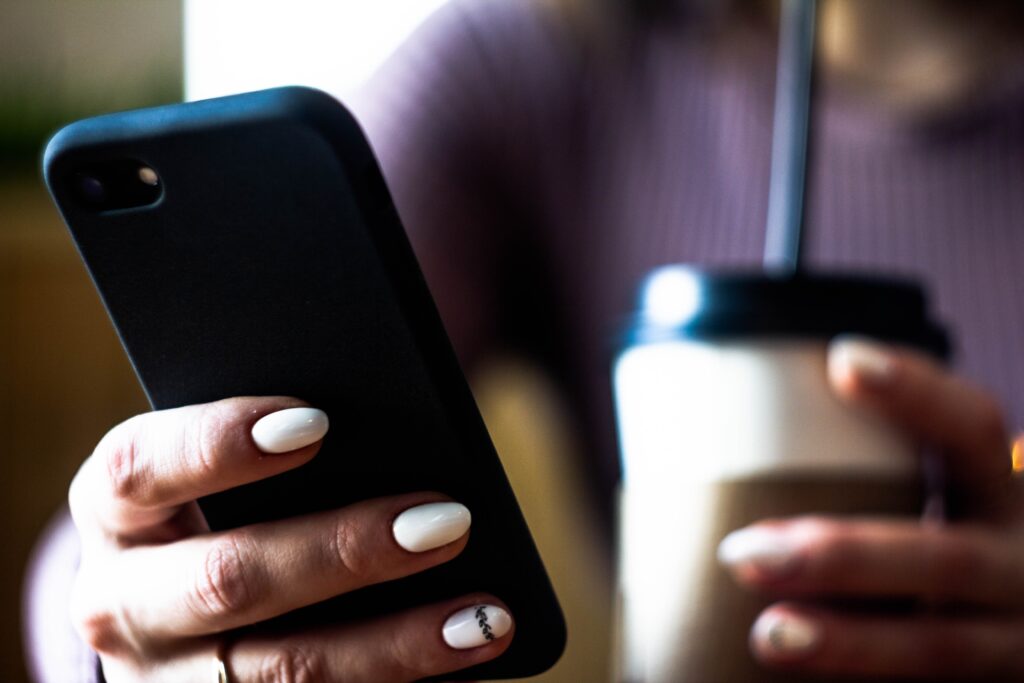 A person with white, manicured nails holds a smartphone in one hand and a takeaway cup with a straw in the other. The background is blurred, and the focus is on their hands and the objects they are holding.