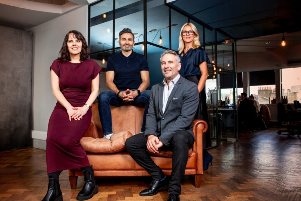 Four people pose in an office with glass walls. Two sit on a brown leather sofa, while two stand behind it. All are dressed in business or smart casual attire and smiling at the camera.
